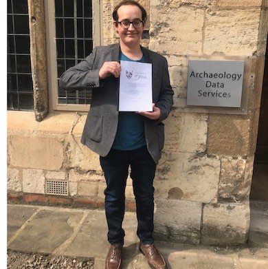 A photo of Alphaeus Talks holding his thesis outside of the Archaeology Data Service.